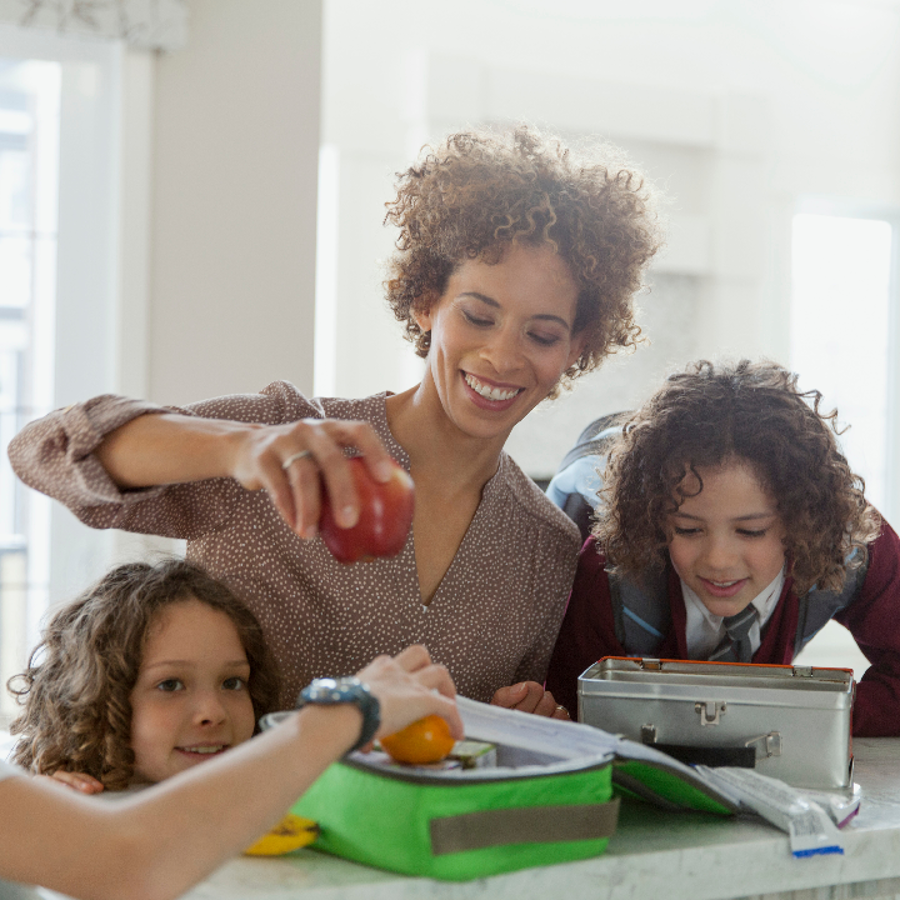 Woman standing in a kitchen surrounded by 3 children whilst she packs an apple into a lunchbox.