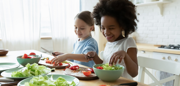 Two young girls sitting at kitchen table smiling and cutting up salad.