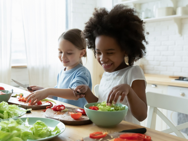 Two young girls sitting at kitchen table smiling and cutting up salad.