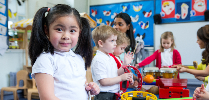 Primary school children in school uniform standing around a table playing with different musical instruments. A young girl in the foreground is staring directly into the camera.