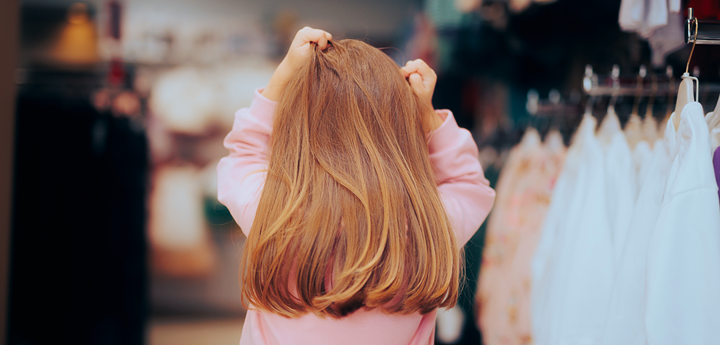 back of a child's head in a clothes shop. She's holding her hands to her head in a stressed gesture