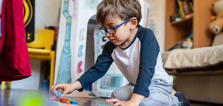 Young boy wearing glasses, kneeling on the floor playing with wooden toys.