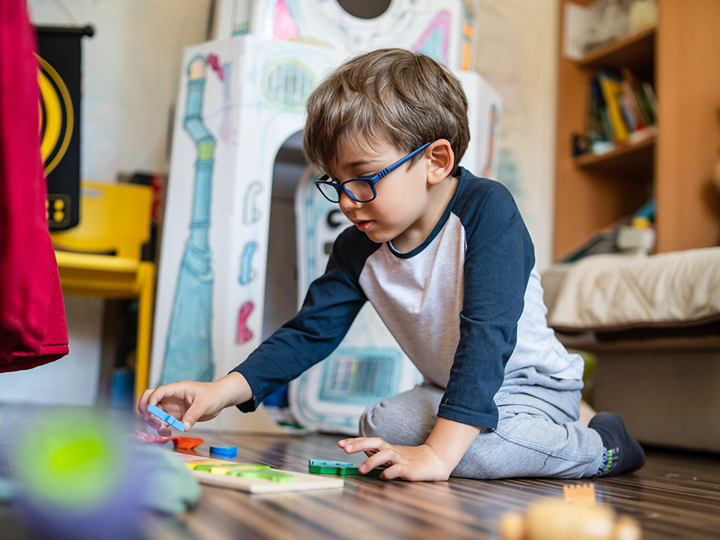 Young boy wearing glasses, kneeling on the floor playing with wooden toys.