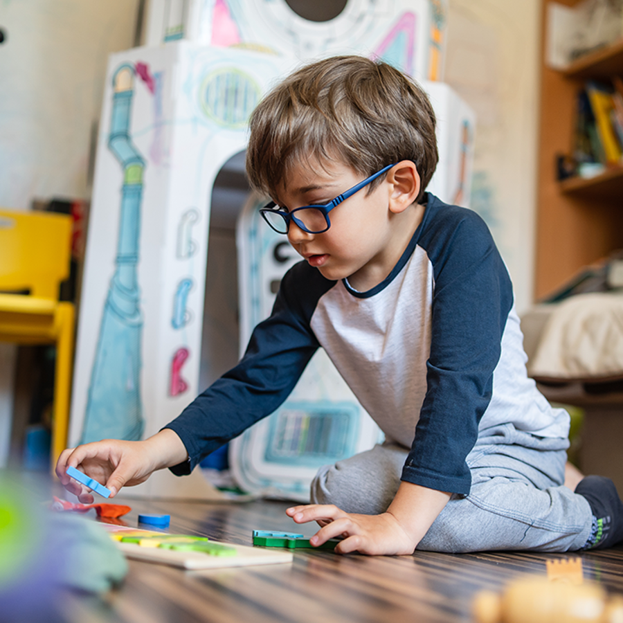 Young boy wearing glasses, kneeling on the floor playing with wooden toys.