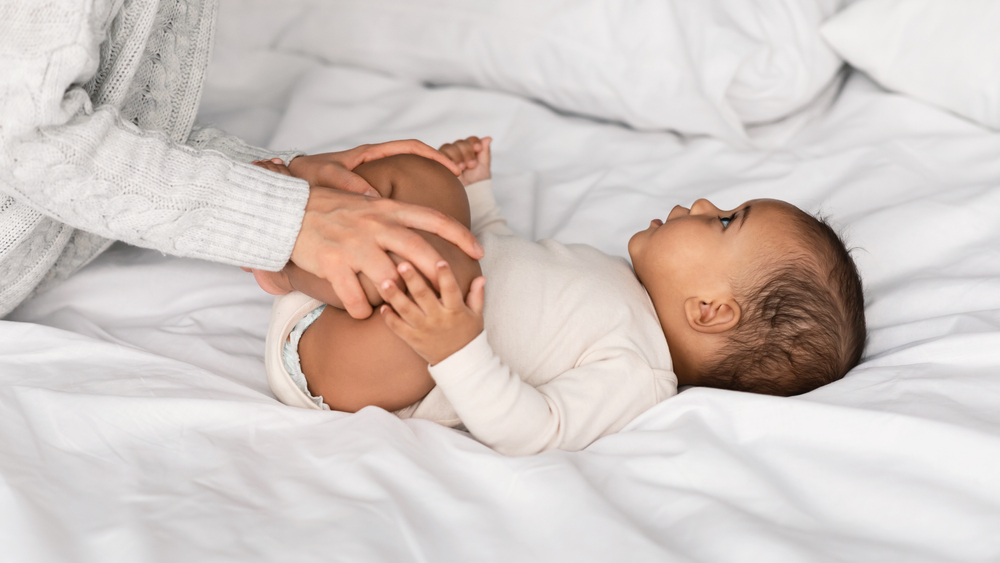 Baby lying on their back on a bed with their mum gently moving their legs to help with their constipation.