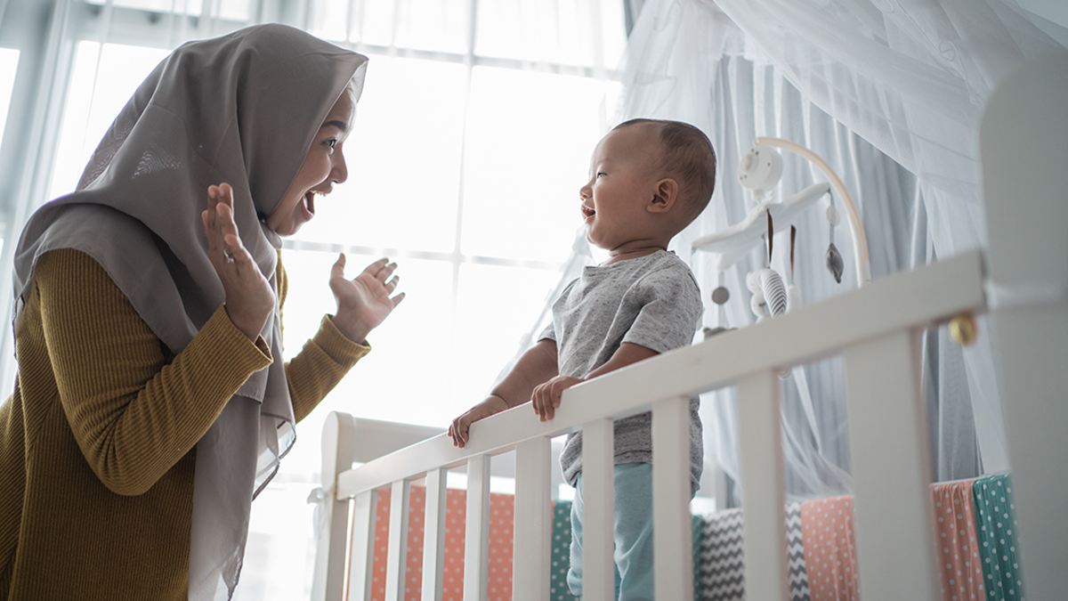 A woman playing peek a boo with their toddler. The toddler is standing up in a cot and the woman is standing next to the cot.