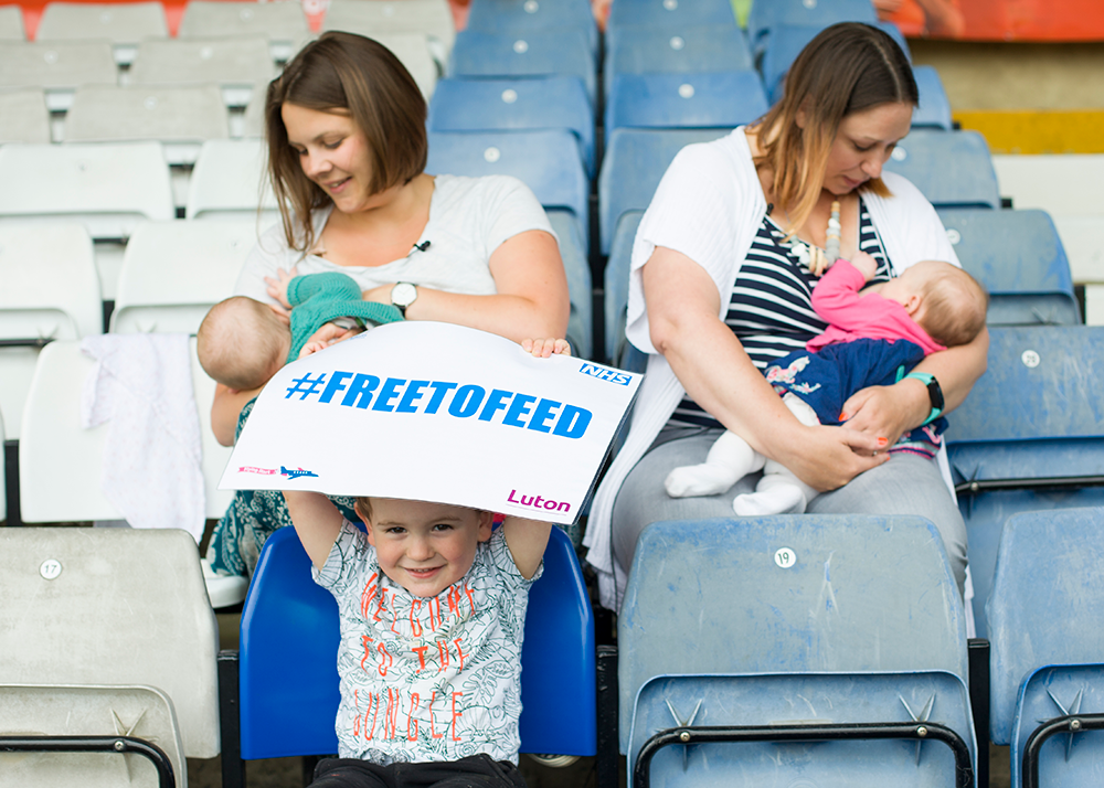 Two Mums Breasteeding Whilst Sitting Next To Each Other In A Football Stadium. On The Chair In Front A Little Boy Is Sitting And Holding A 'Free To Feed' Sign About His Head