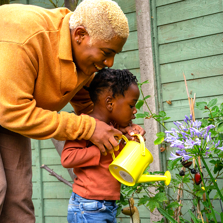 adult and child using a bright yellow watering can to water a plant outside