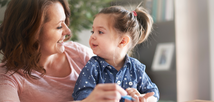 Toddler girl sitting on adult woman's lap at a table. The toddler and woman are both holding a crayon and smiling at one another whilst drawing on a piece of paper in front of them.