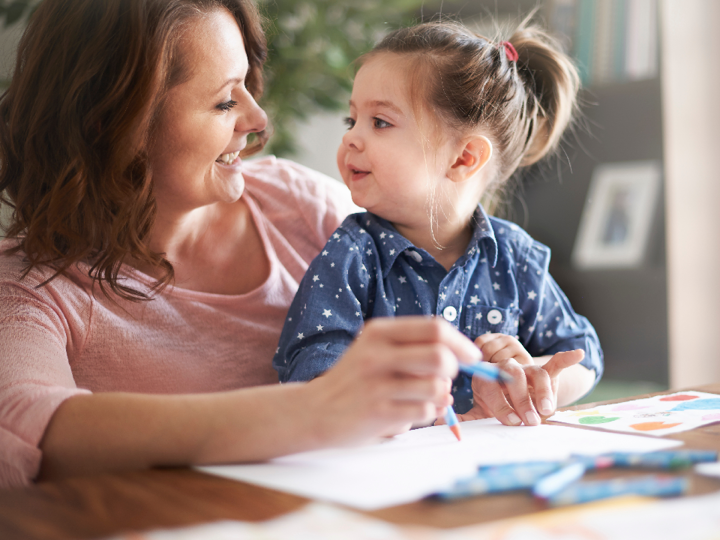 Toddler girl sitting on adult woman's lap at a table. The toddler and woman are both holding a crayon and smiling at one another whilst drawing on a piece of paper in front of them.