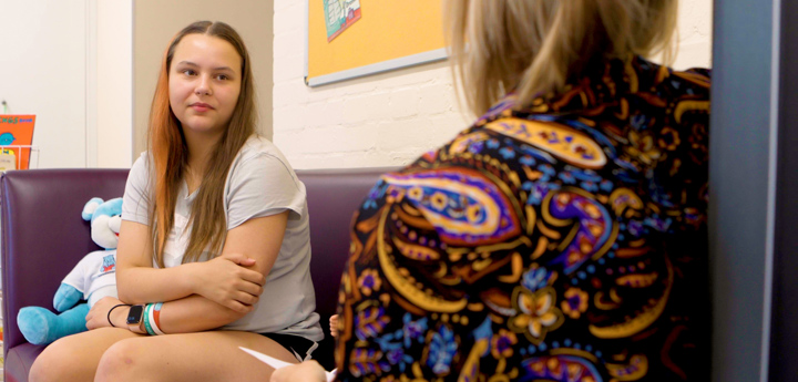 Young woman sat with arms crossed on a sofa, speaking to a woman in the foreground