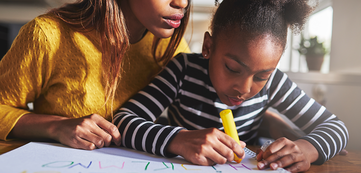 Young girl writing in coloured pens on a large sheet of paper. An adult woman is sitting behind the girl leaning over her shoulder and looking at what the girl is writing.