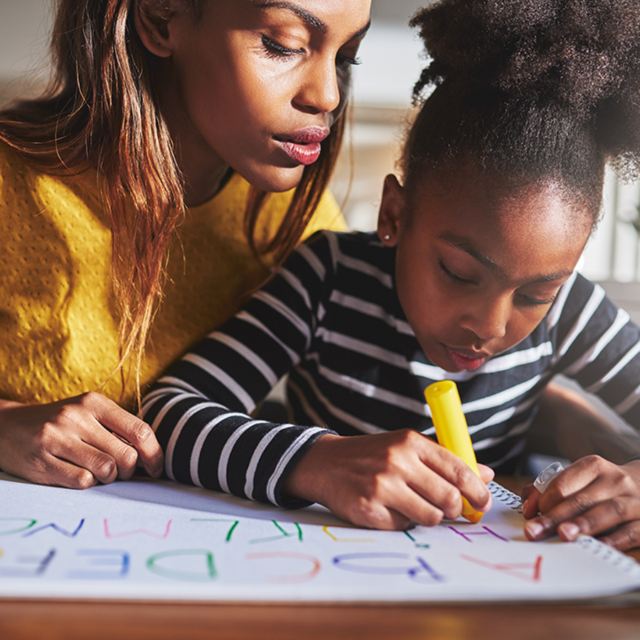 Young girl writing in coloured pens on a large sheet of paper. An adult woman is sitting behind the girl leaning over her shoulder and looking at what the girl is writing.