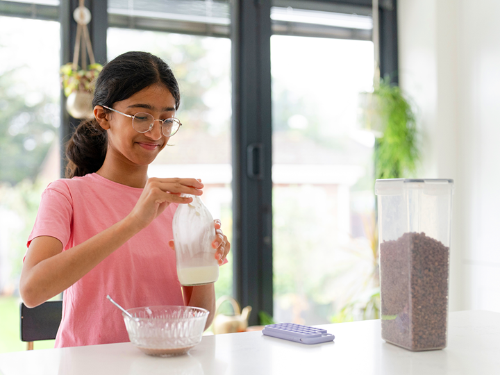 Teenage girl wearing glasses in kitchen holding a milk bottle over a bowl of cereal.