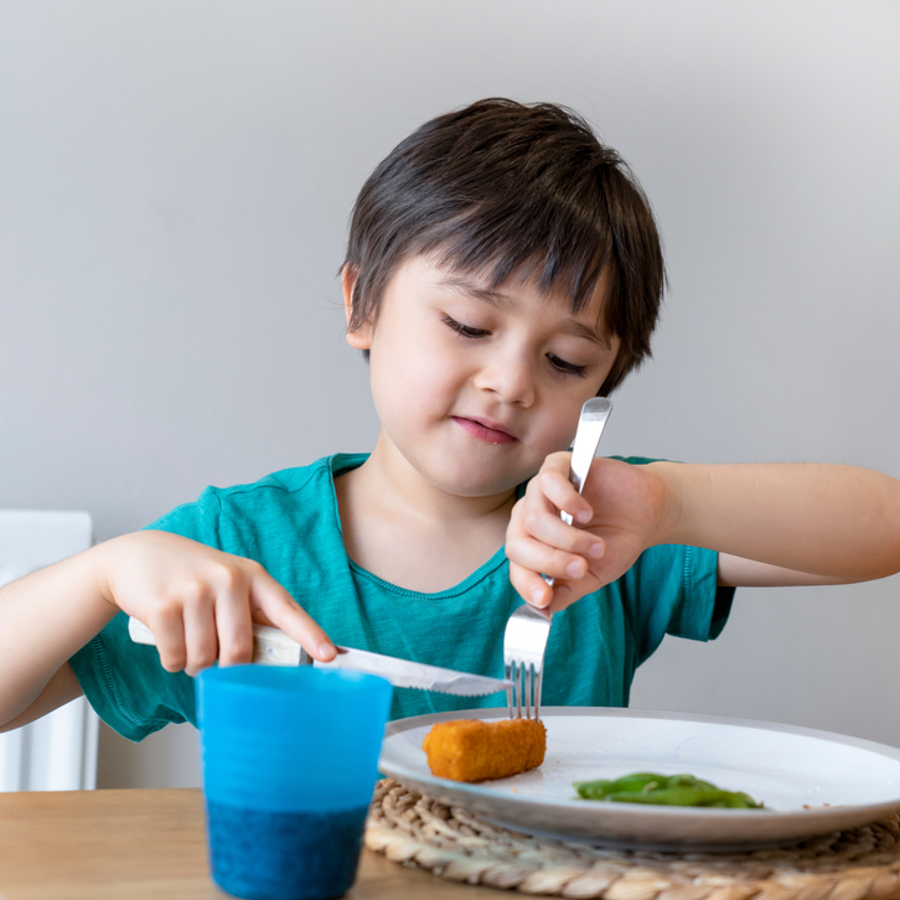 Child sitting at a table. They are cutting a fish finger using a knife and fork.