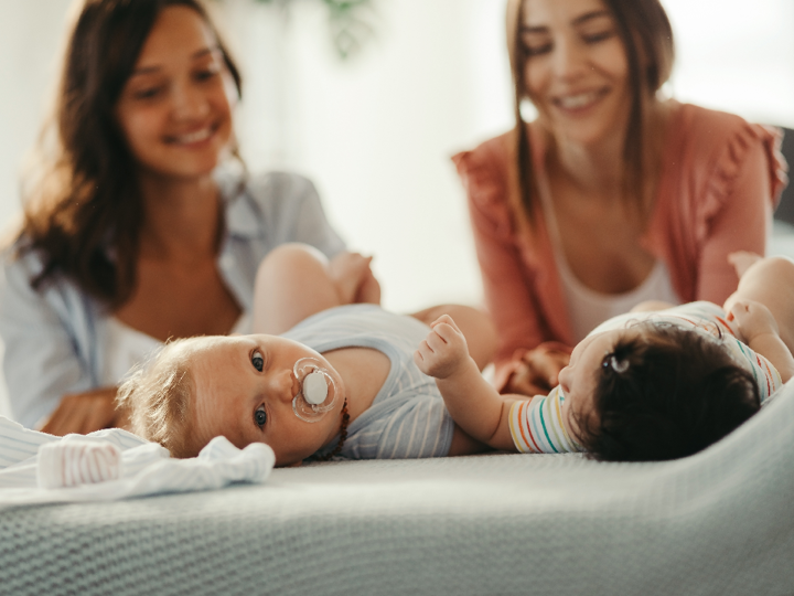 Two women kneeling down in front of their babies. The two babies are lying on their back. One baby is looking at the other, whilst the other baby is looking at the camera.