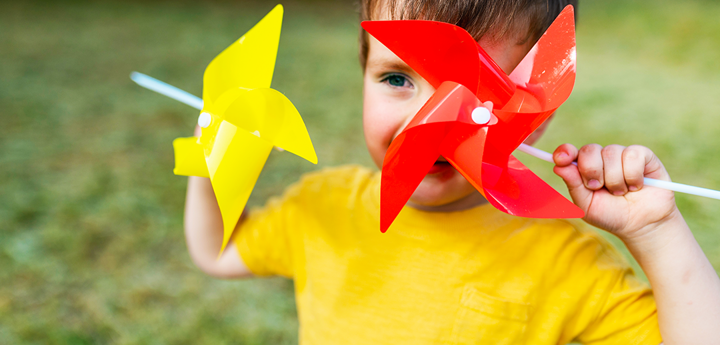 Boy holding 2 pinwheels to his face and smiling. 1 eye is visible through the pinwheels.