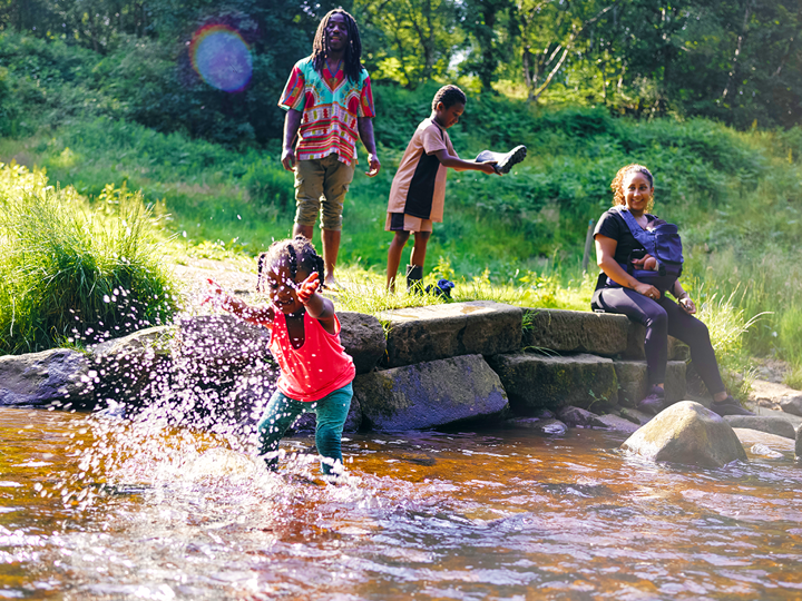 Child playing in a river, adults looking on smiling, one adult has a baby on their chest and a child in the background looking at the inside of their wellies