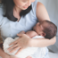 A woman sitting down and smiling whilst looking down at the baby she is holding, which is wrapped in a blanket, in her arms