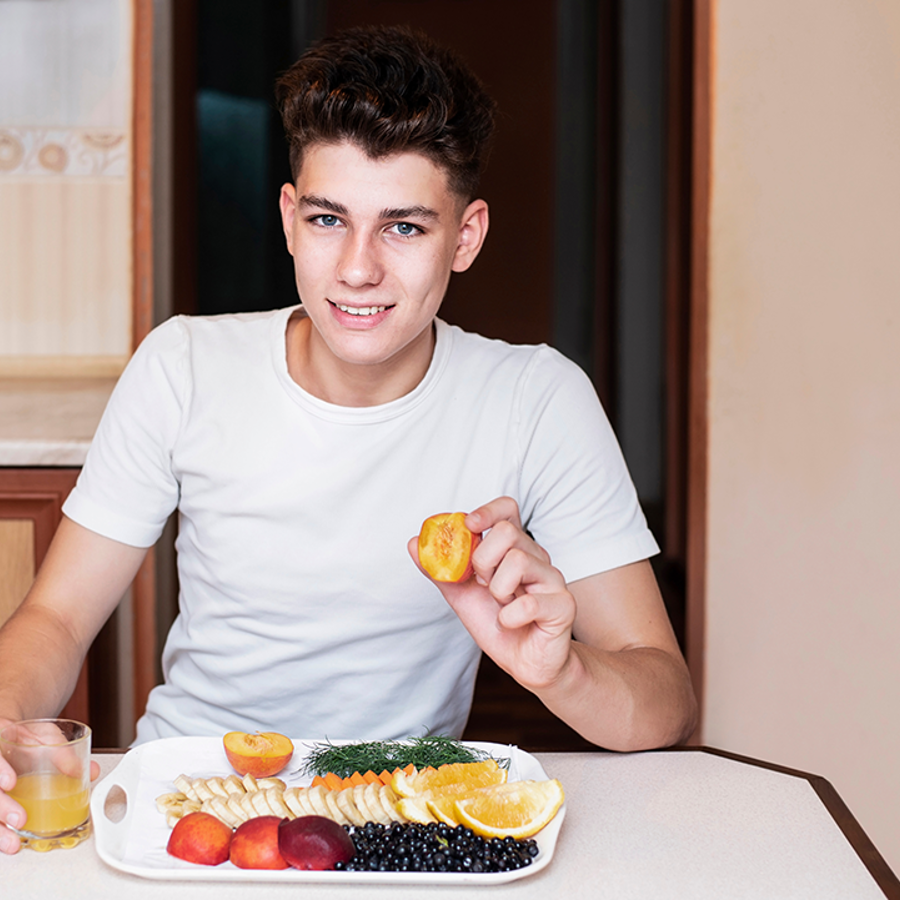 Teenage boy sitting at a table smiling holding a piece of fruit in one hand and a glass of juice in the other. On the table is a plate of fruit in front of him.