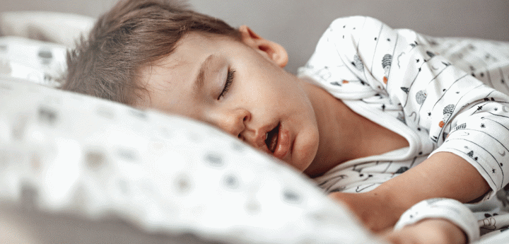 A boy sleeping in his bed on his side with his head laying against a pillow and arms outstretched.