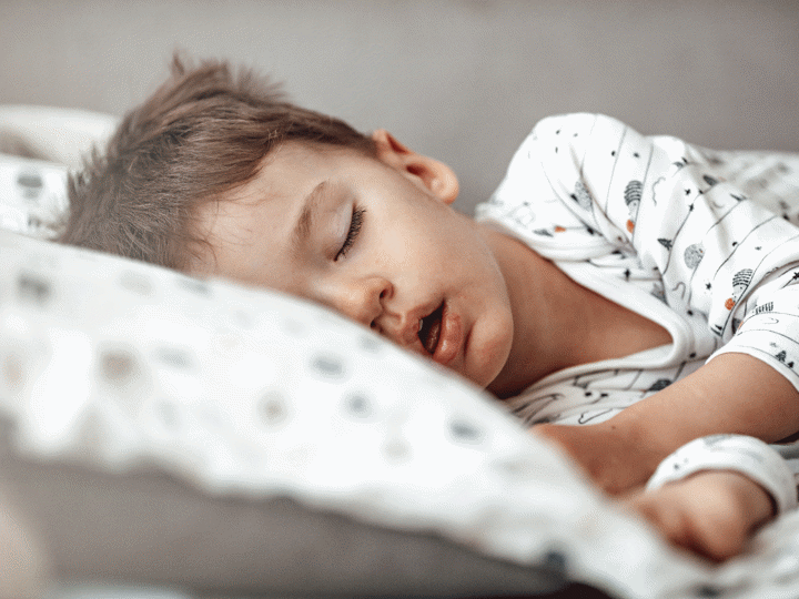 A boy sleeping in his bed on his side with his head laying against a pillow and arms outstretched.