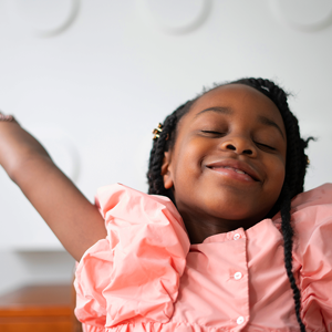 A child smiling with her eyes closed. She's wearing a pink dress with her arms raised above her head gracefully.