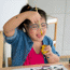 Young girl with glasses painting, sitting at a table dipping a paintbrush into a paint pot and smiling.