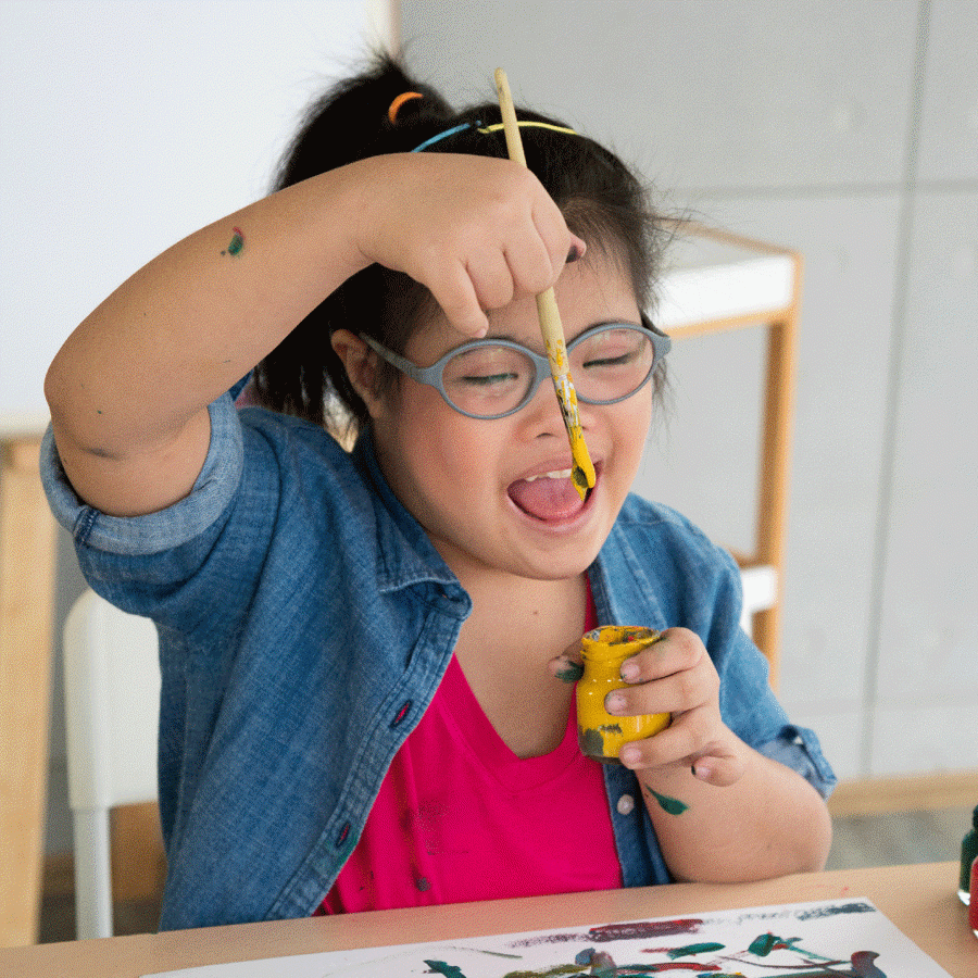 Young girl with glasses painting, sitting at a table dipping a paintbrush into a paint pot and smiling.