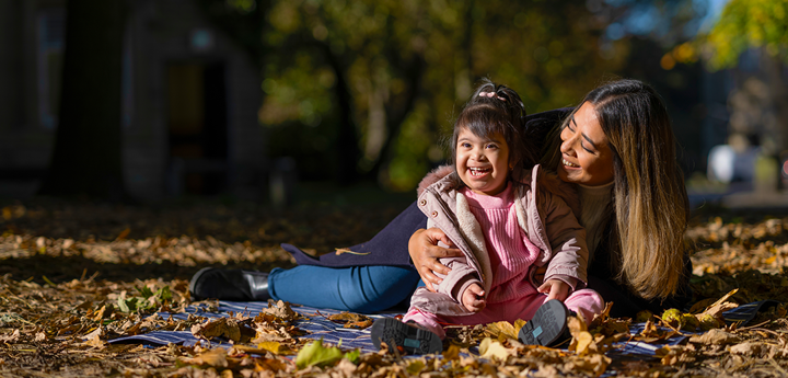 Down's syndrome child and parent laughing and sitting on picnic blanket in autumn with leaves around them.