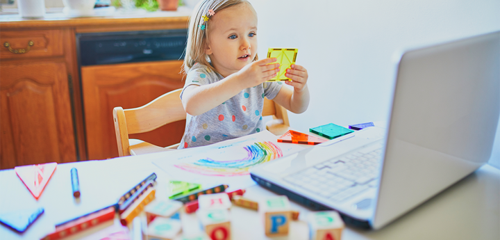 A young girl playing with shapes sitting at a kitchen table in front of a laptop.