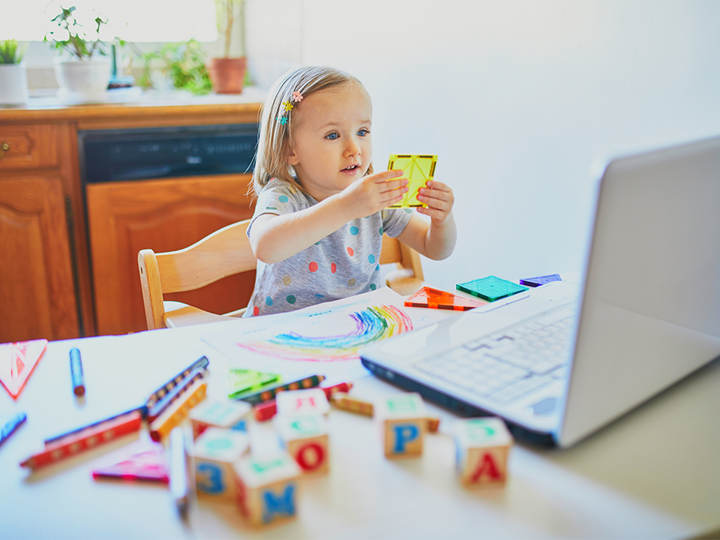 A young girl playing with shapes sitting at a kitchen table in front of a laptop.