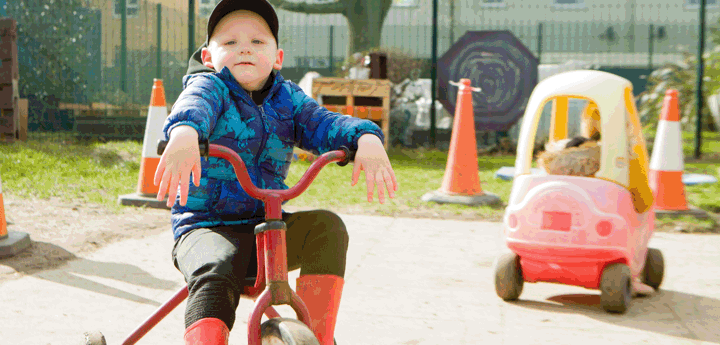 Boy sitting on tricycle with red and yellow car in background