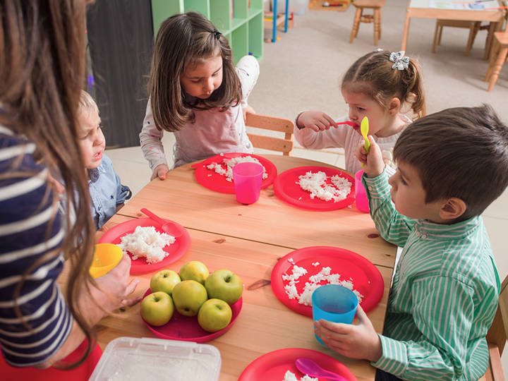 Group of 4 young children sitting round a table eating food from plastic plates.