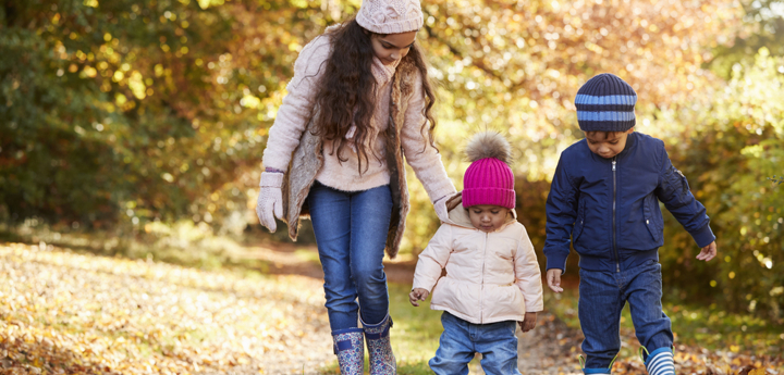 A young girl and two toddlers walking through the woods on a cold day wearing welly boots, coats and hats.