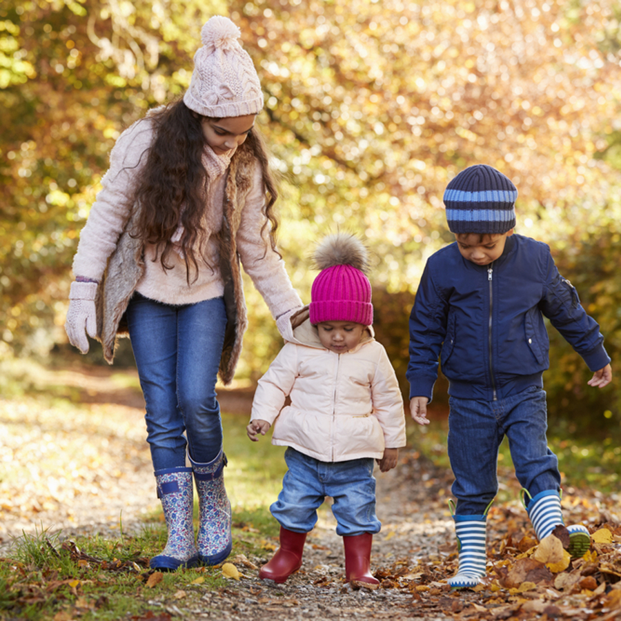 A young girl and two toddlers walking through the woods on a cold day wearing welly boots, coats and hats.