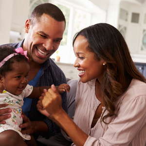 Mum and dad sitting on sofa laughing and smiling with a happy baby girl sitting on the dad's knee