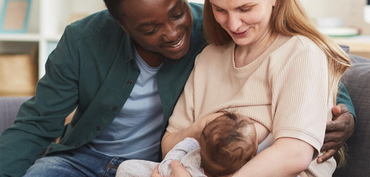 Mum sitting on sofa breastfeeding her baby with the dad sitting next to her with his arm round her. 