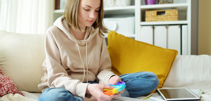 teenage girl on bed, legs crossed playing with fidget toy on bed with ipad and notebook in front of her