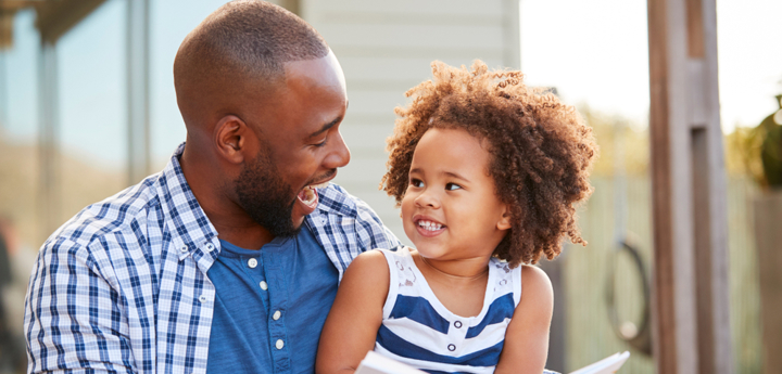 Young girl sitting on dad's knee reading a book together and laughing.