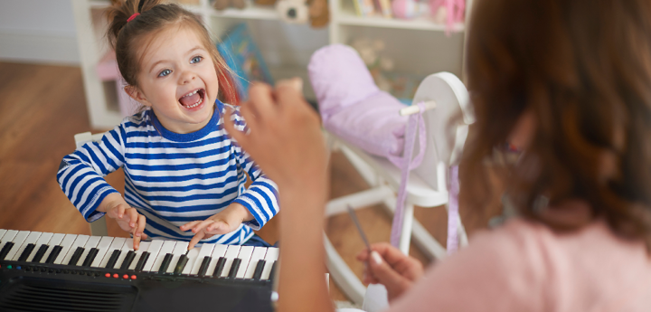 Young girl smiling whilst pushing keys on a keyboard. A woman in the foreground is facing the girl and conducting. 