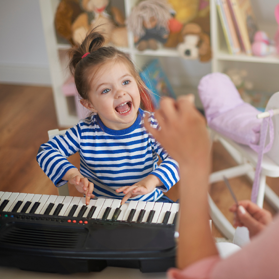 Young girl smiling whilst pushing keys on a keyboard. A woman in the foreground is facing the girl and conducting. 