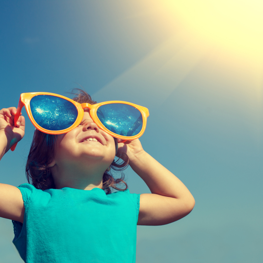 Young girl smiling whilst wearing oversized yellow sunglasses looking up at the sky.