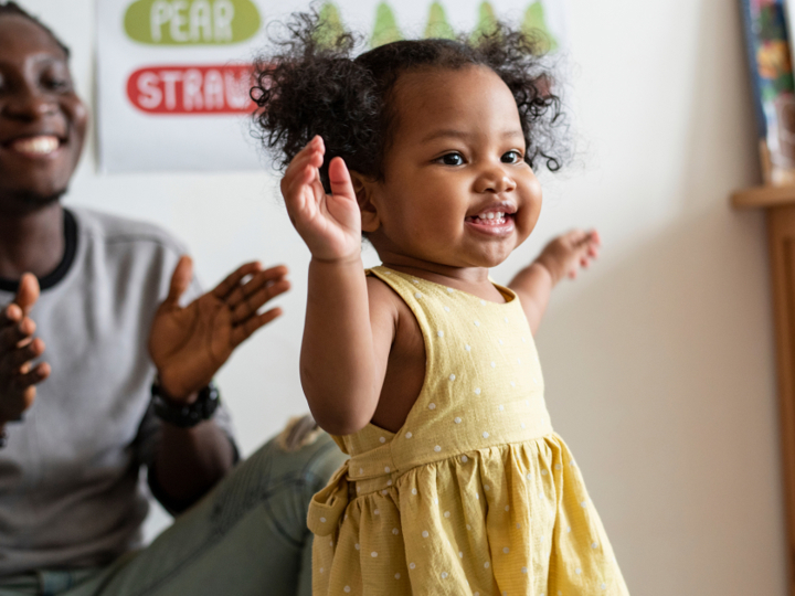 Toddler taking first steps with smiling adult in background