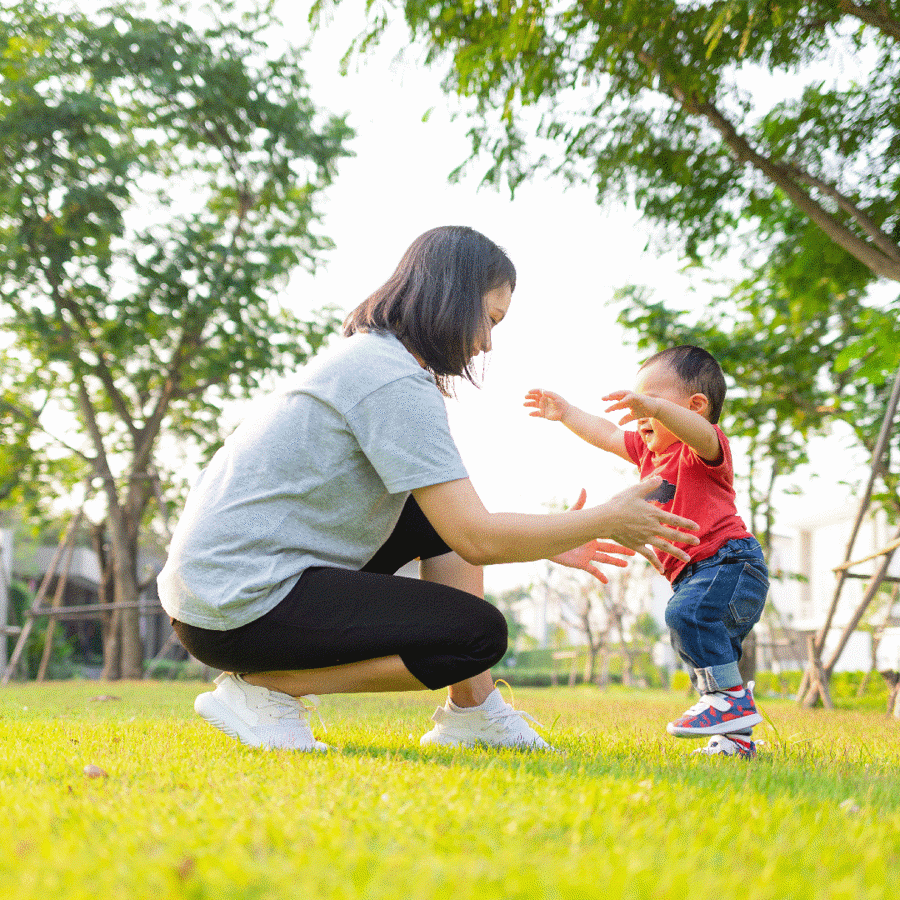 A mum bending down on the grass, teaching their child to walk outside. 