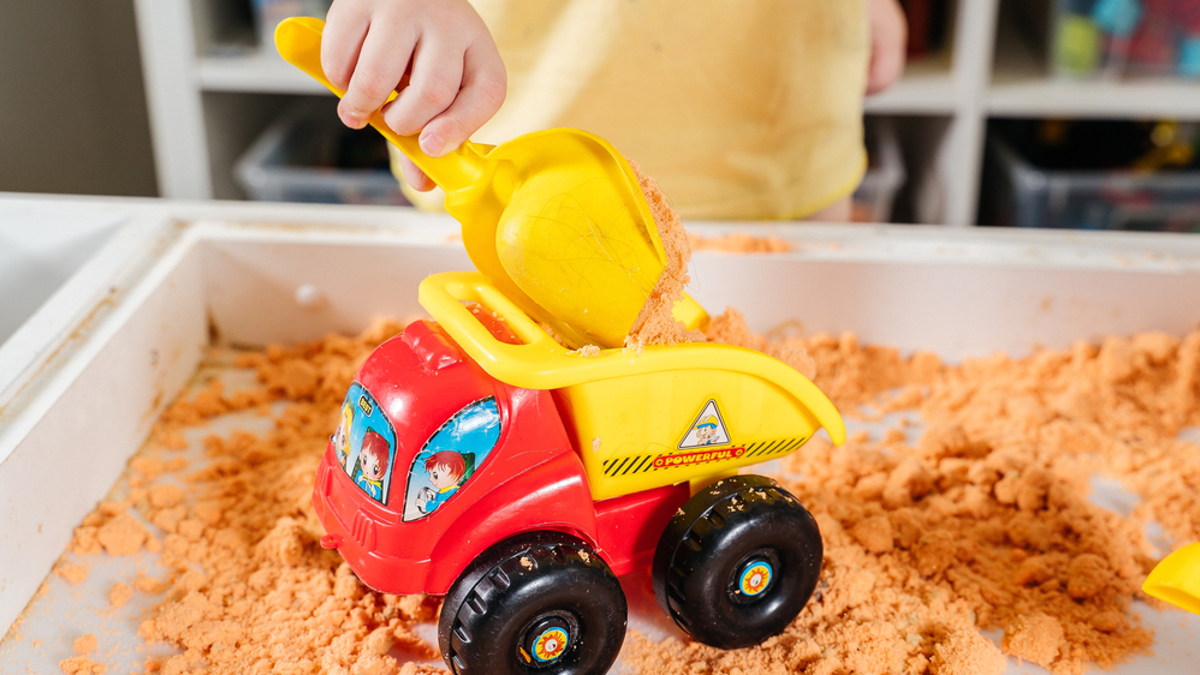 Child playing scooping sand using yellow plastic trowel into plastic truck.