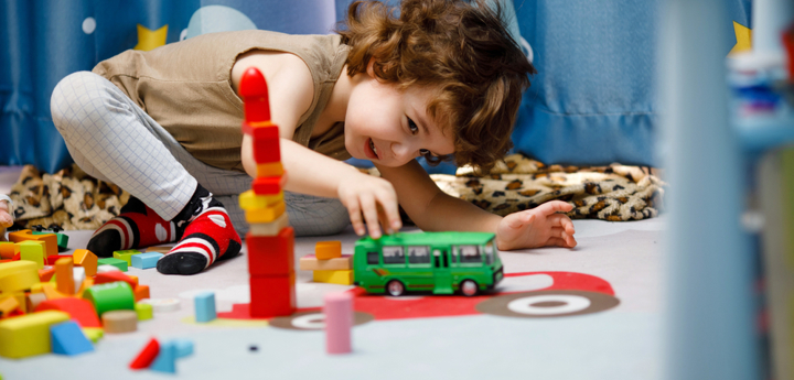 Young child sitting on the floor. They are playing with plastic blocks and a green toy car.