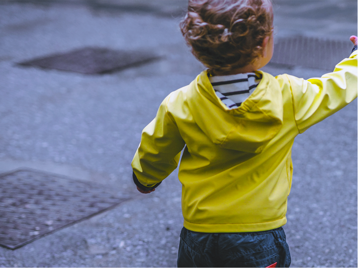 Boy toddler standing near the road in a fluorescent yellow jacket reaching his hand out.