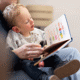 Boy looking at book whilst sitting in his parents lap. The parent is holding the book reading it to the young boy.