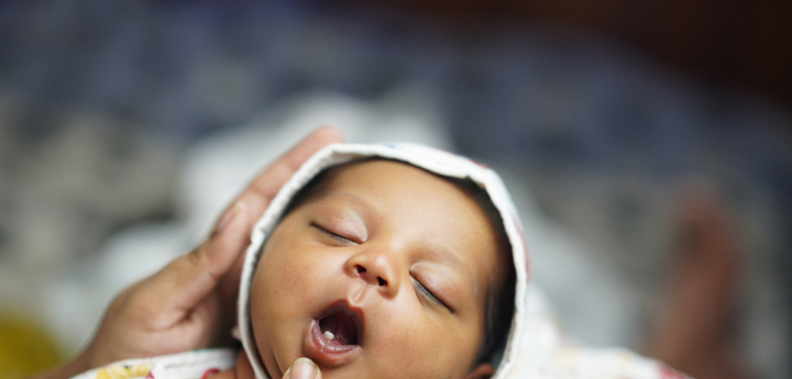 Sleeping newborn baby being held with an adults finger gently pulling down on their lower lip to reveal the baby's first tooth.